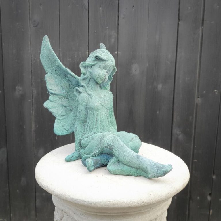 A wing adorned fairy sitting with legs splayed out in front of her. Situated on a pedestal in front of a British garden fence