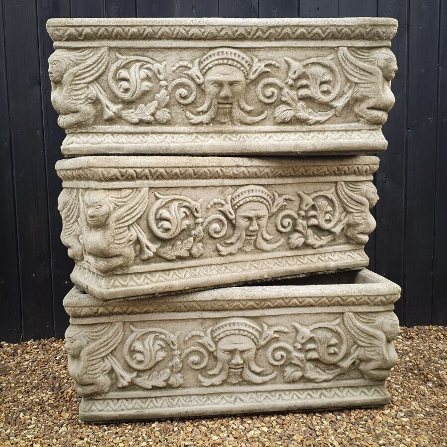 3 large toughs laid on top of each other with intricate decoration featuring carved faces and birds. Situated by the fence of a British garden