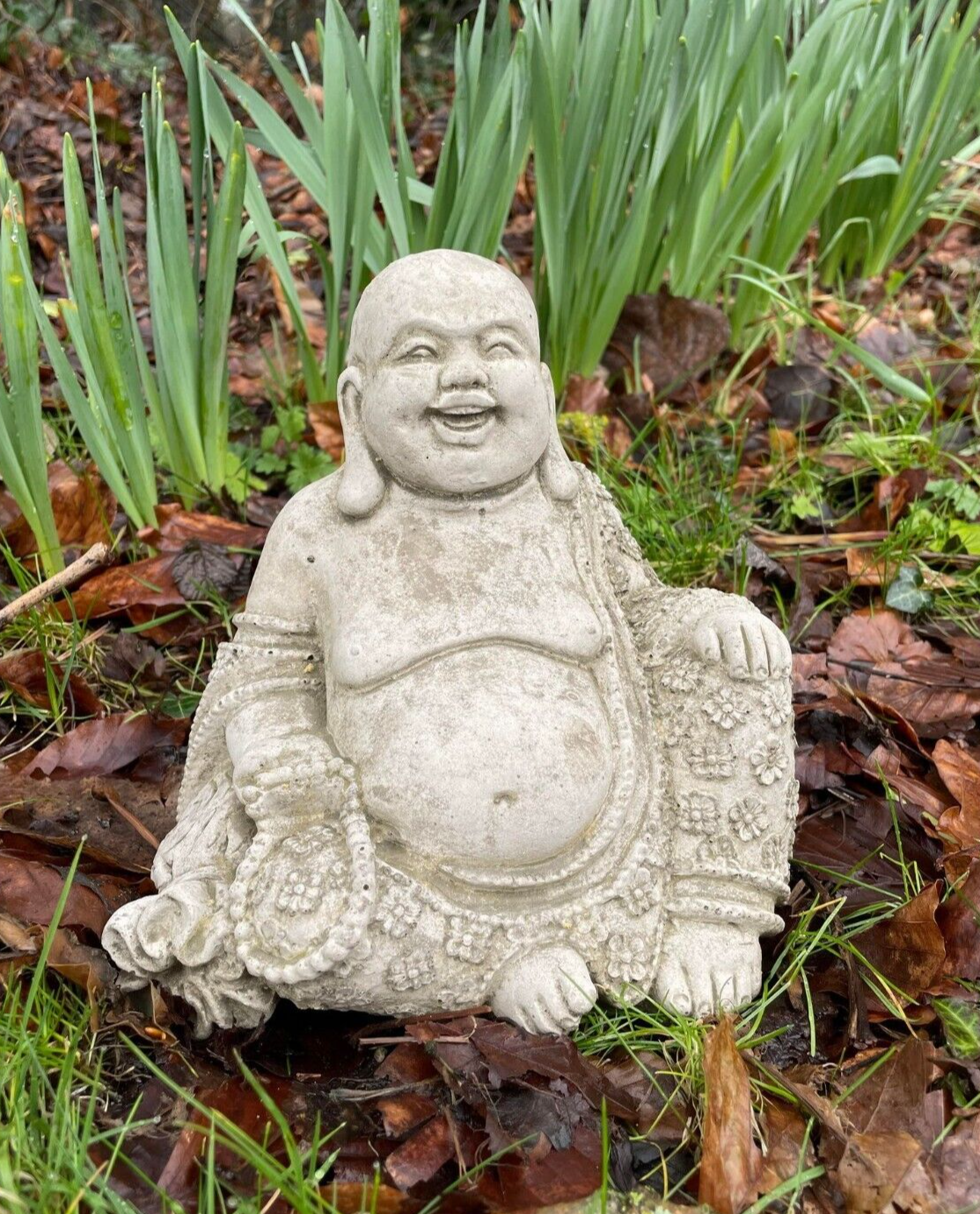 A large bellied Buddha with stretched ear-lobes and decorative garments. Wearing a large grin, sitting in the foliage of a British garden