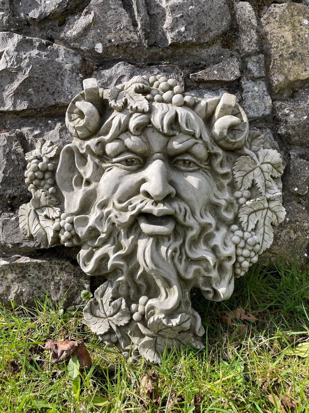 A large bachus head hanging plaque adorned with berries and leaves with flowing beard and rams horns. Situated leaning on the wall of a British garden with green grass in the foreground.
