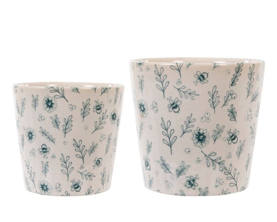Two round ceramic plant pots of different sizes in white finish. Adorned with bumblebee and floral design. Situated on a white studio background