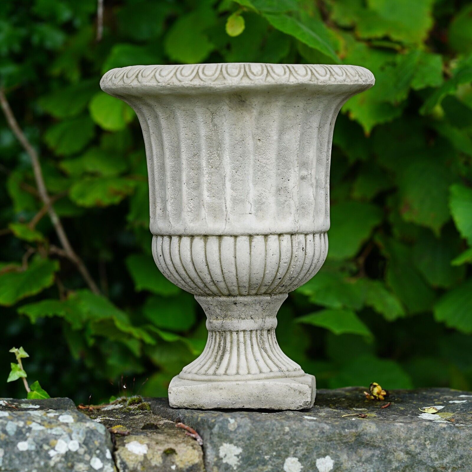 A Victorian style stone urn with intricate pattern design. Situated on the garden wall of a British garden with green foliage in the background