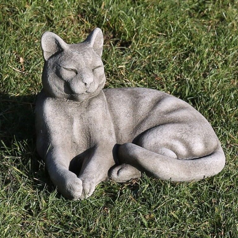An elegant stone cat with curled tail and sleepy eyes, laying amongst the grass of a British garden
